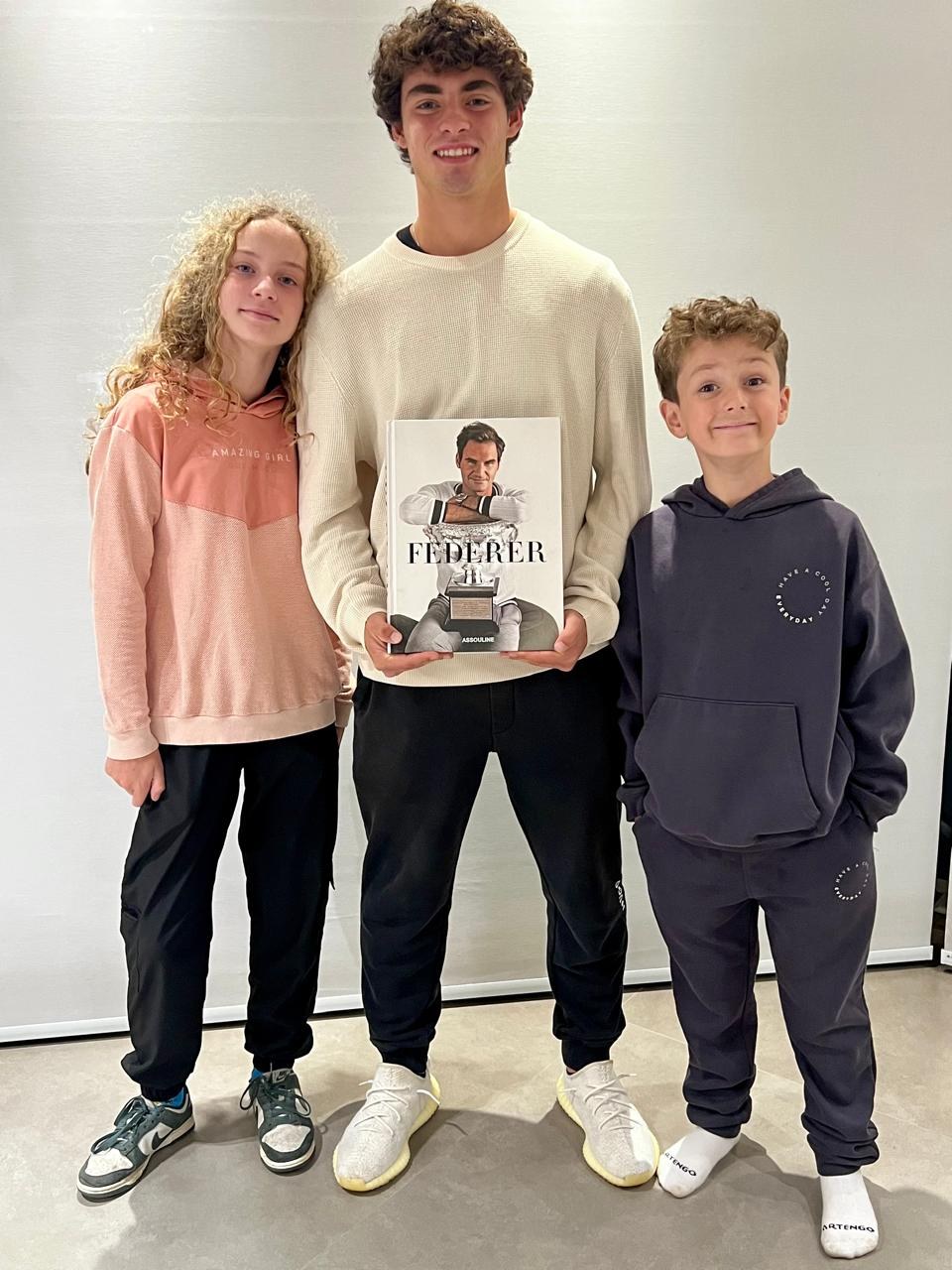 Os três filhos do Eder lado a lado, cada um de uma altura e de um jeito: a do meio de cabelo cacheado, o mais velho no centro segurando um livro sobre o Federer, o caçula de moletom escuro.