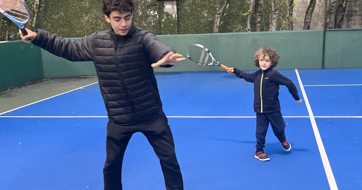 O filho mais velho, adolescente, mostra o movimento do forehand na quadra azul, e o caçula, pequeno, imita atrás dele com a raquete na mão.